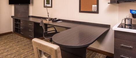 A hotel room with a dark wood desk, a flat-screen TV, a corkboard, a small kitchenette, and a wicker chair.