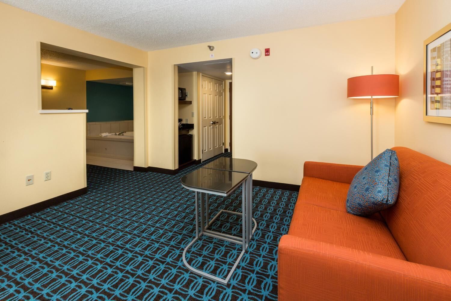 Una habitación de hotel con alfombra azul estampada, sofá naranja con almohada azul, una mesa de vidrio, una lámpara de pie roja y una puerta que da a un baño.