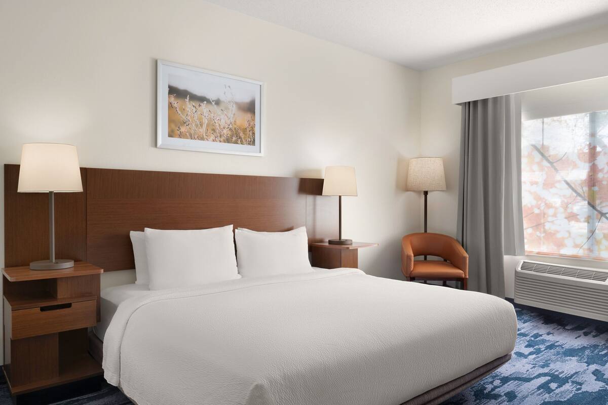 Una camera d'albergo con un letto grande, due comodini, una sedia, un quadro incorniciato appeso al muro e una finestra con tende.
