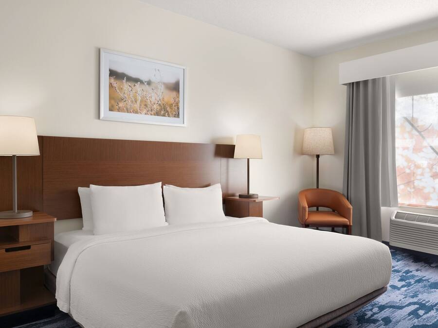 Habitación de hotel con una cama grande, dos mesitas de noche, una silla, un cuadro enmarcado en la pared y una ventana con cortinas.