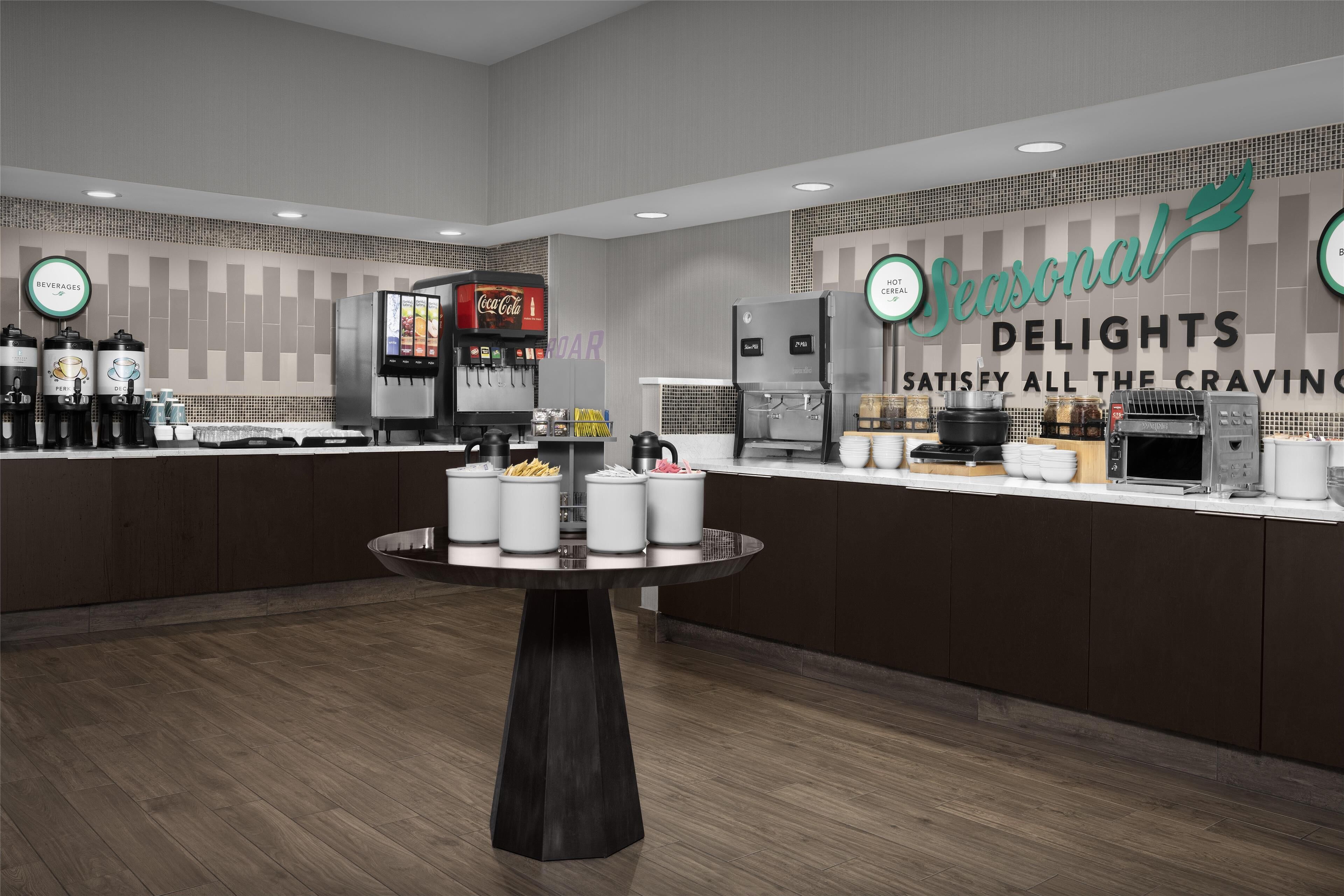 Ein modernes Café mit einem runden Tisch in der Mitte, einer Coca-Cola-Maschine, einer Kaffeemaschine und einem Schild mit der Aufschrift 'Seasonal Delights'.