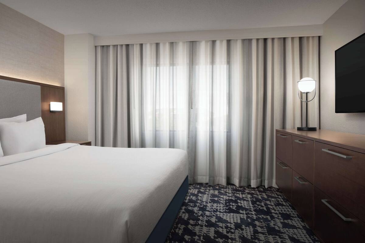 Une chambre d’hôtel avec un grand lit, une lampe de chevet, un téléviseur à écran plat, une commode et un tapis à motifs.