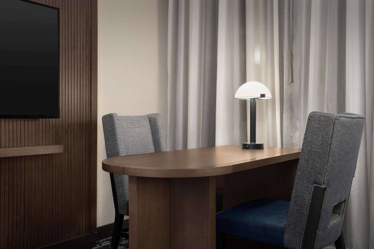 Une chambre d’hôtel avec une table à manger en bois, deux chaises grises à revêtement, un canapé bleu, une lampe sur pied et une grande fenêtre avec des rideaux.