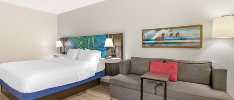 A hotel room with a bed, a sofa, a nightstand with lamps, a painting on the wall, and a wooden floor.