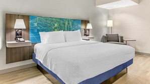 A modern hotel room with a large bed, a desk, two chairs, a nightstand with a lamp, and a wall art piece.