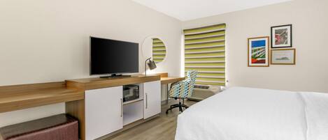 A hotel room with a bed, a desk with a flat-screen TV, a microwave, a chair, and a window with blinds.