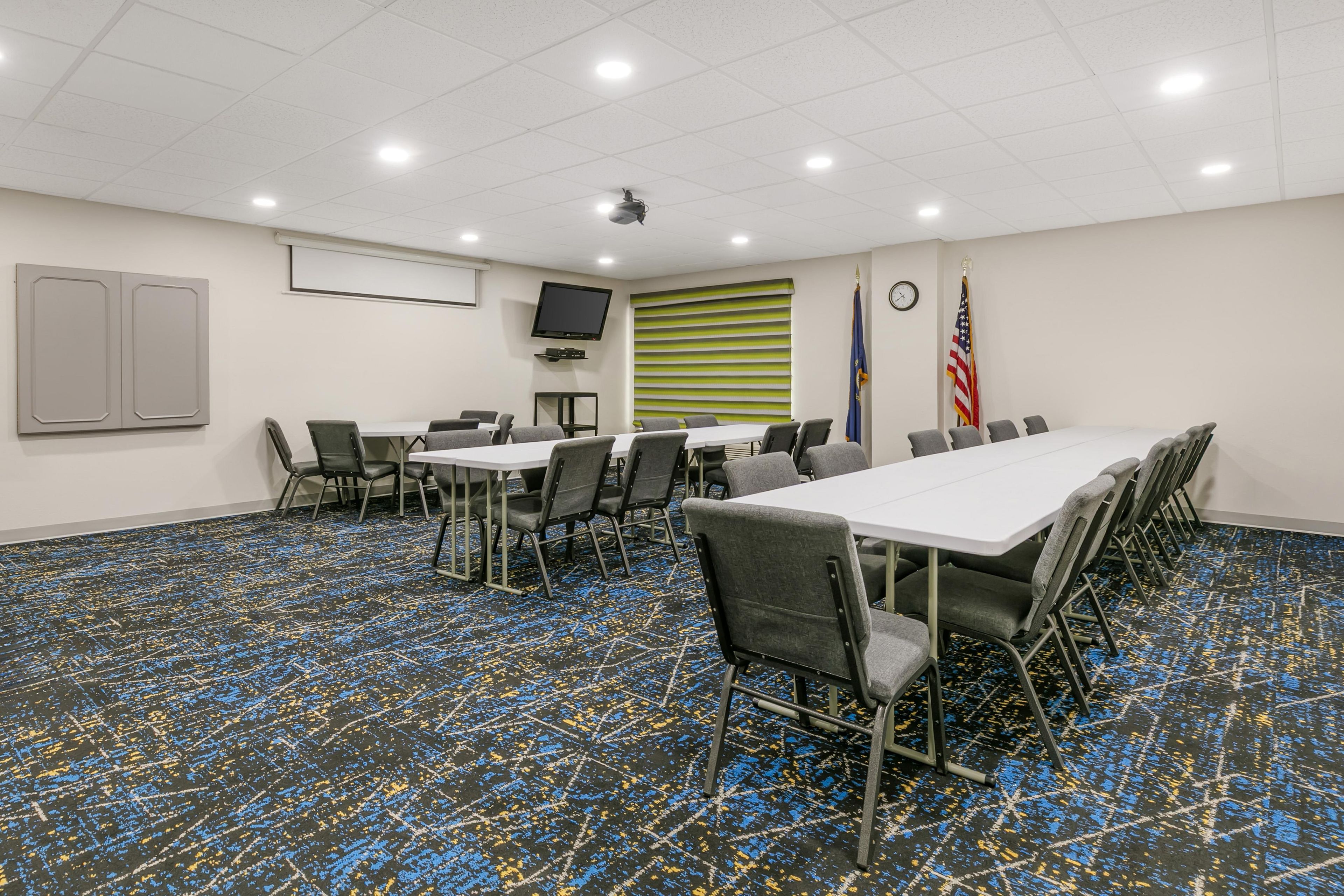 A conference room with a long table, chairs, a projector, flags, and a clock on the wall.