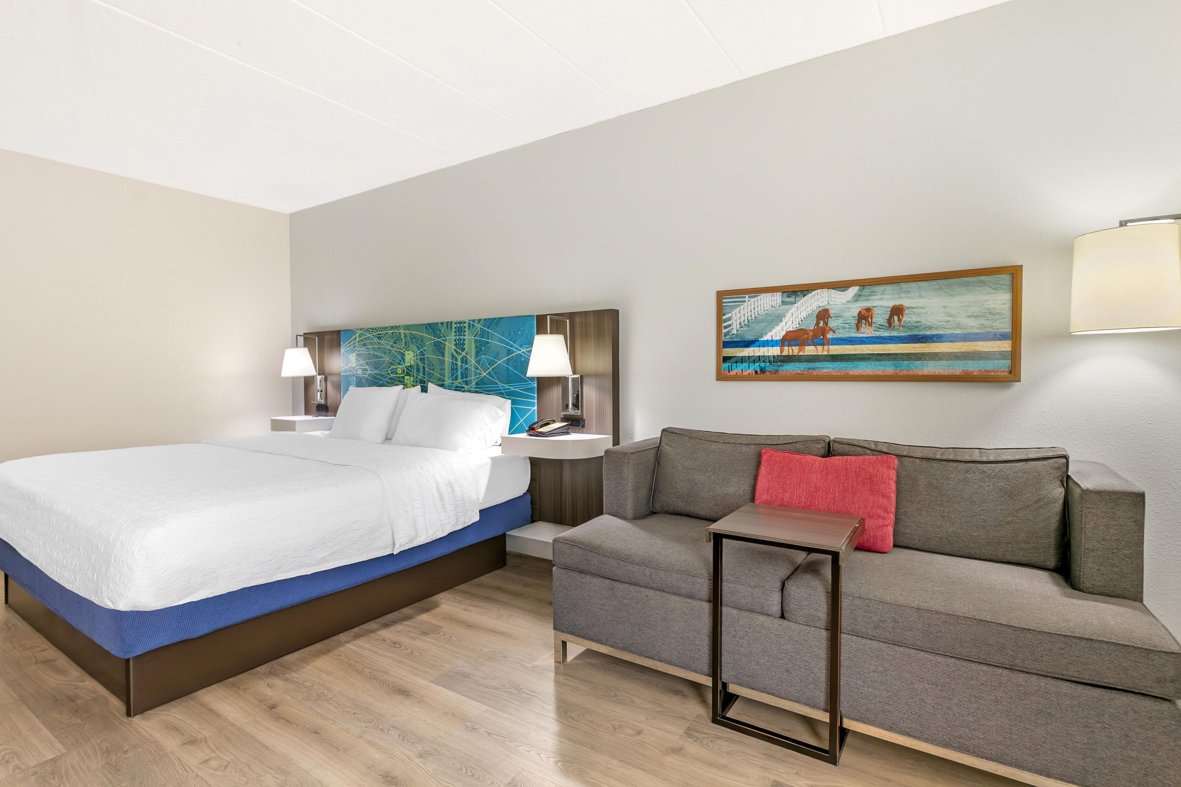 A hotel room with a bed, a sofa, a nightstand with lamps, a painting on the wall, and a wooden floor.