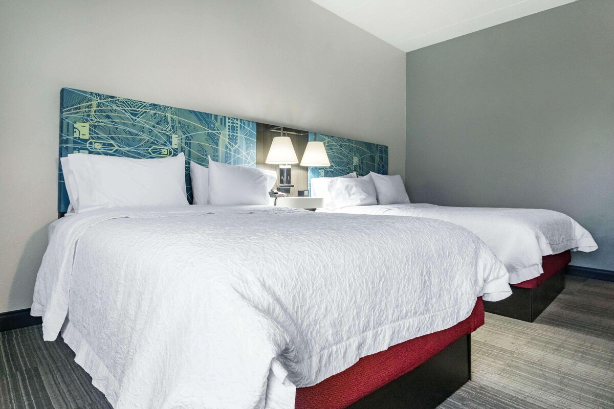 A hotel room with two beds, a headboard with a cityscape design, and a lamp between the beds.