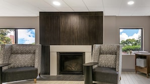 A modern room with a fireplace, two patterned armchairs, a small table, and a view of trees through large windows.