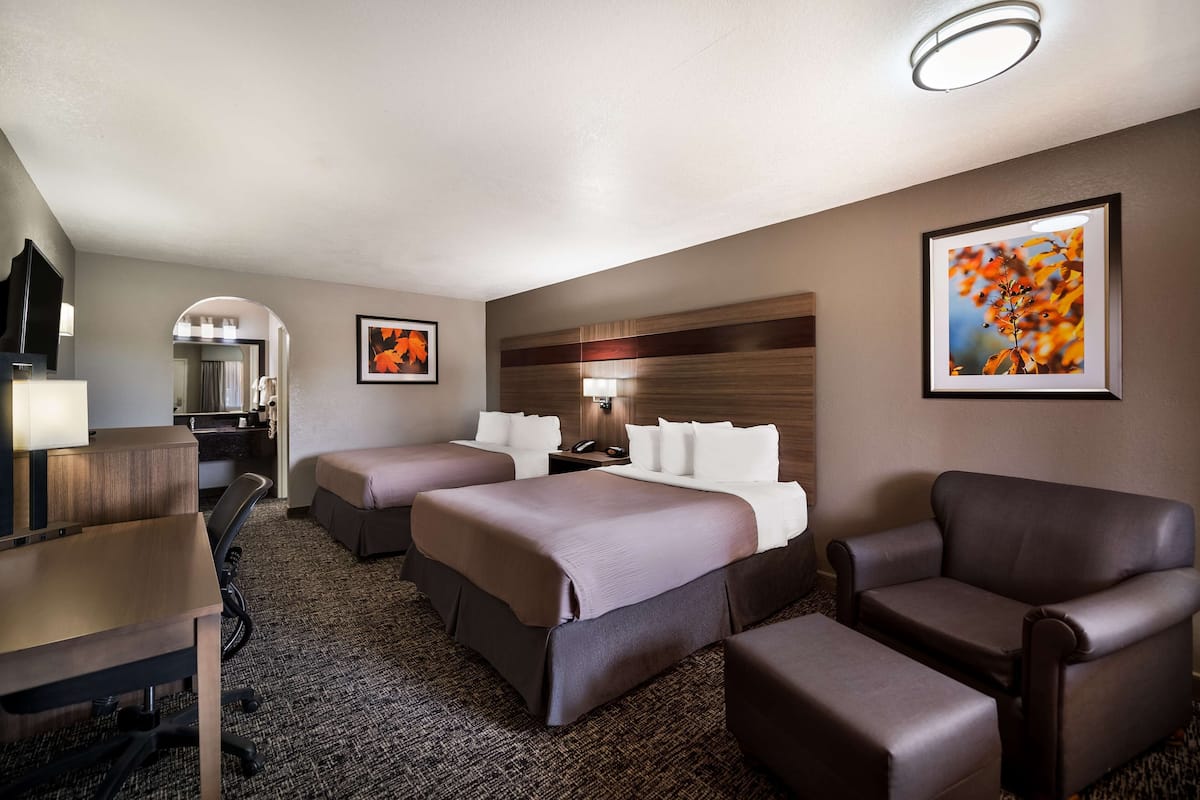 Ein Hotelzimmer mit zwei Betten, einem Schreibtisch, einem Stuhl und einer Couch. An der Wand befinden sich ein Spiegel, ein Bilderrahmen und eine Lampe.