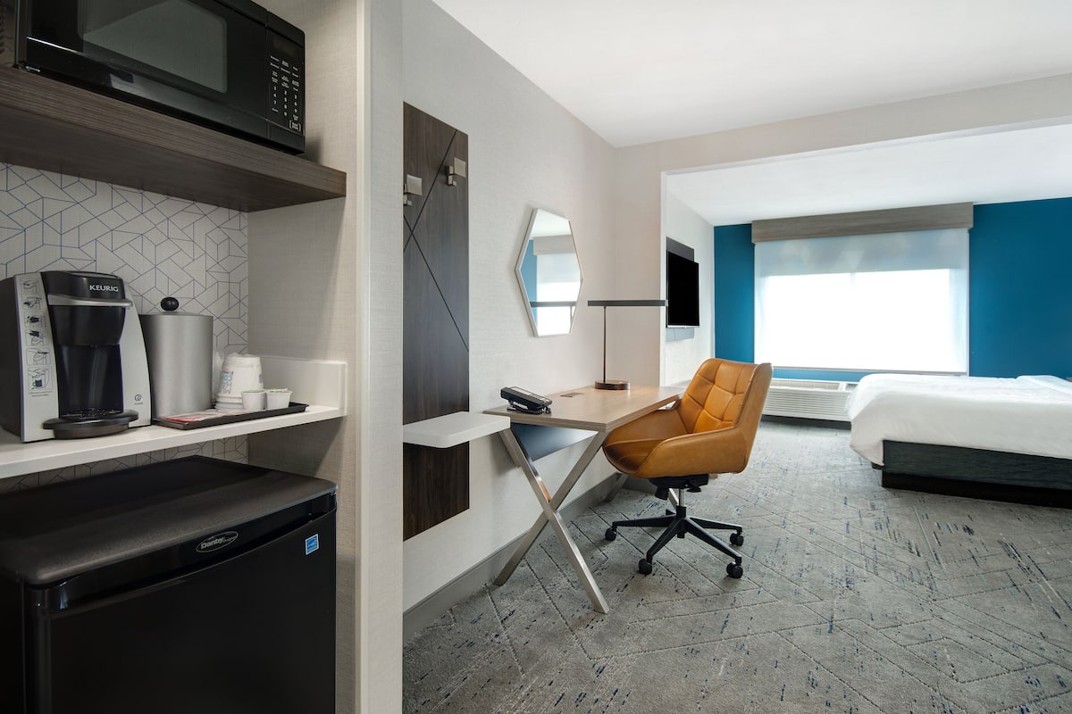 Habitación de hotel con cama, escritorio con silla, cafetera, microondas y refrigerador pequeño.