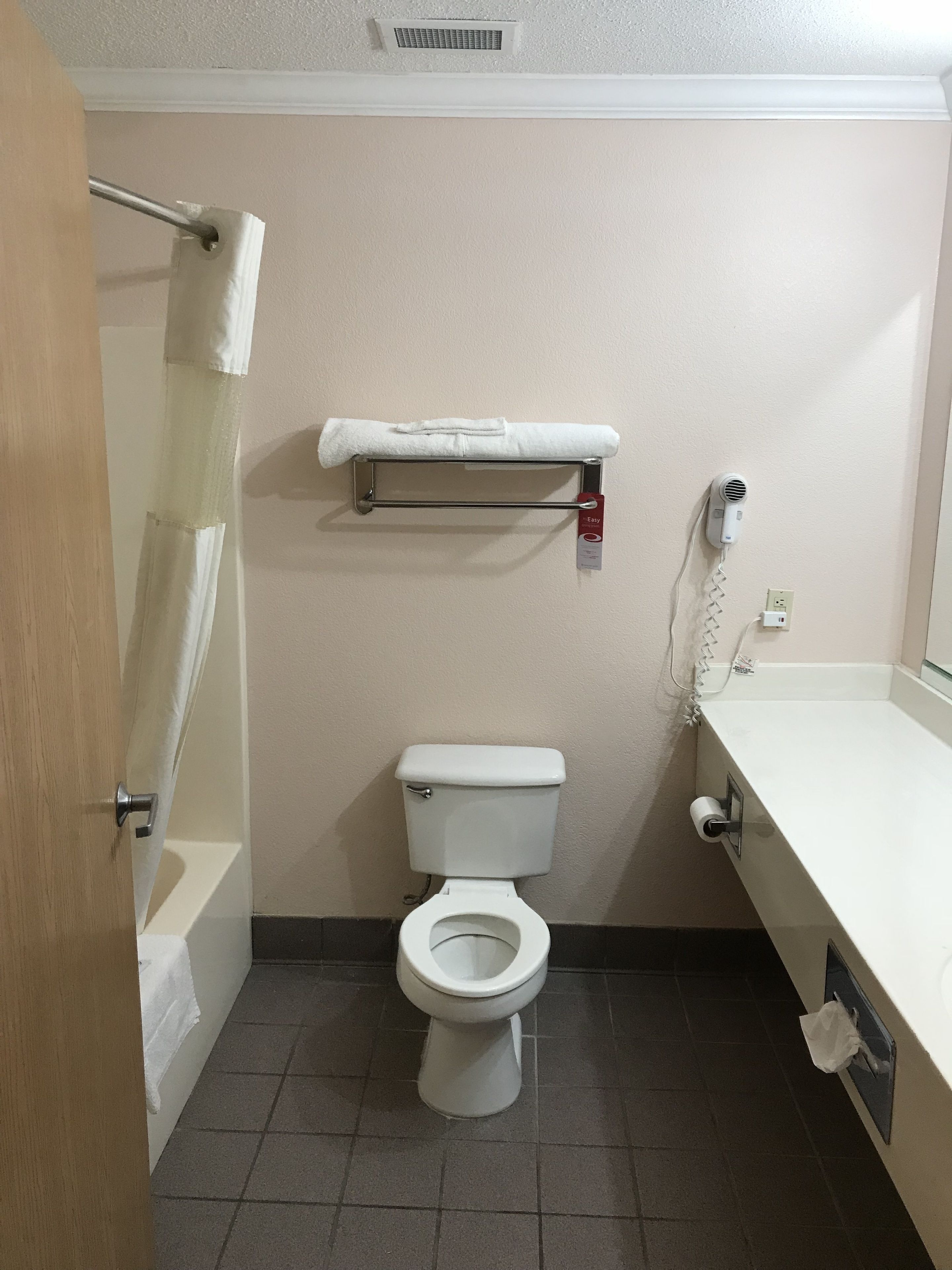 Une salle de bain équipée de toilettes, d’un porte-serviettes, d’un rideau de douche, d’un distributeur de serviettes et d’un sèche-cheveux.