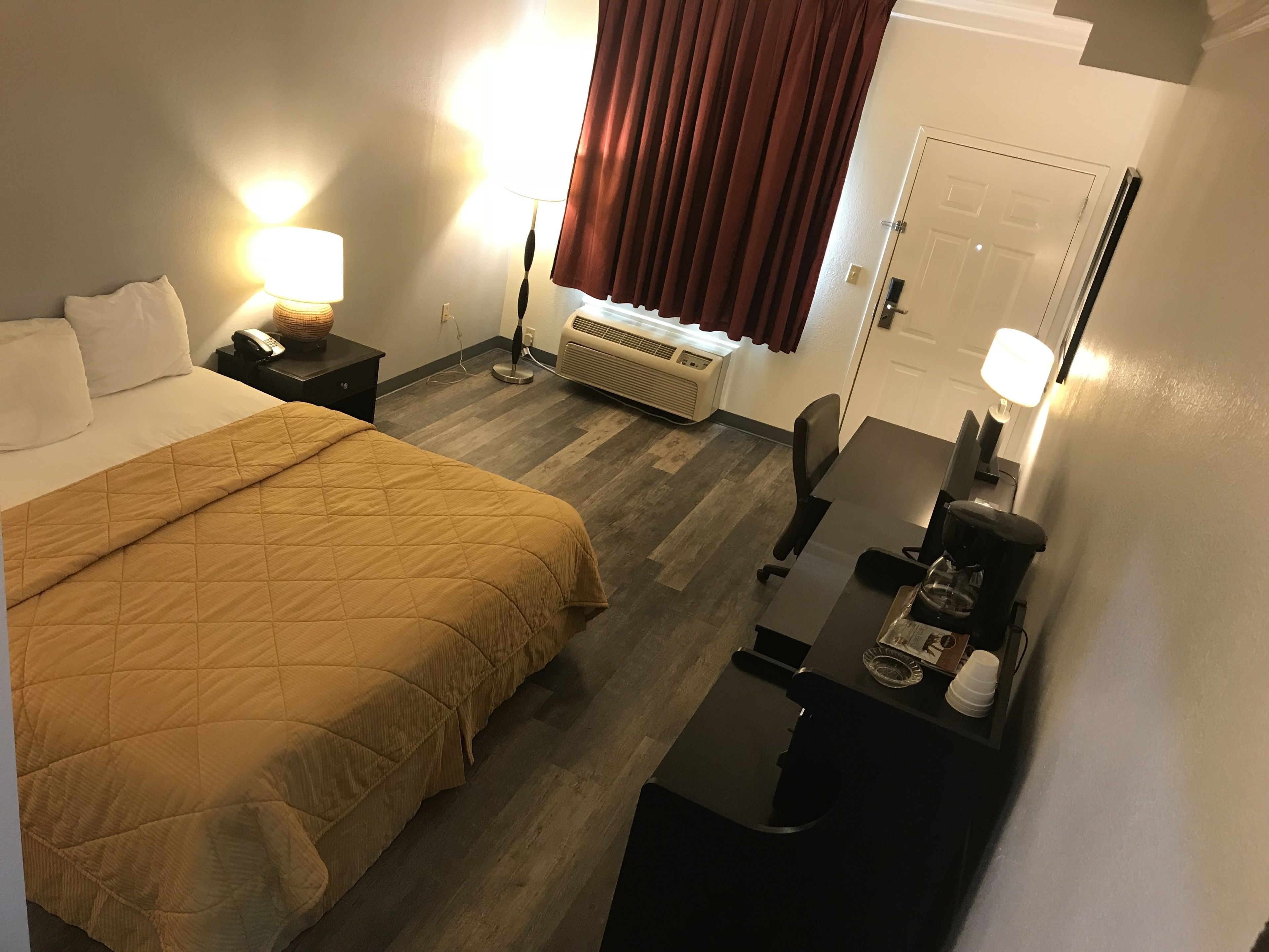 A hotel room with a bed, bedside table, lamp, desk with chair, and an air conditioning unit.