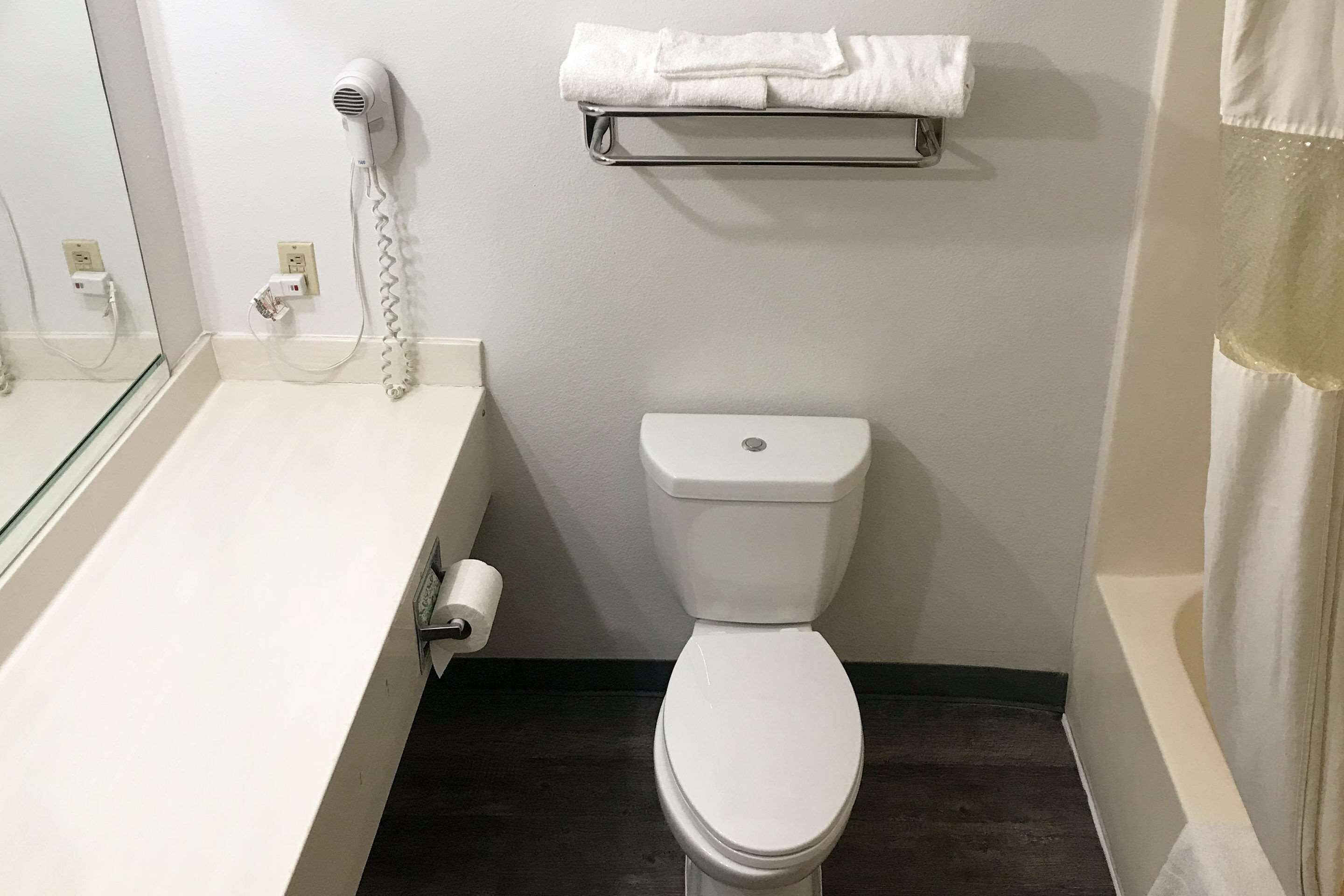 Une salle de bain avec des toilettes blanches, un sèche-cheveux fixé au mur, un porte-serviettes et un support à papier toilette.