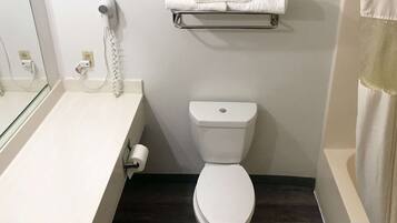 Une salle de bain avec des toilettes blanches, un sèche-cheveux fixé au mur, un porte-serviettes et un support à papier toilette.
