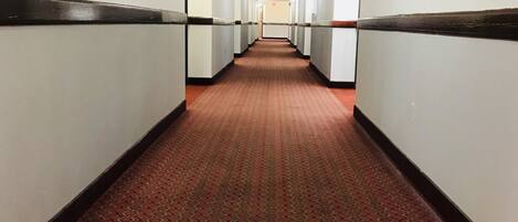 Un long couloir étroit d’hôtel, avec un tapis à motifs, des murs blancs et un panneau de sortie rouge.