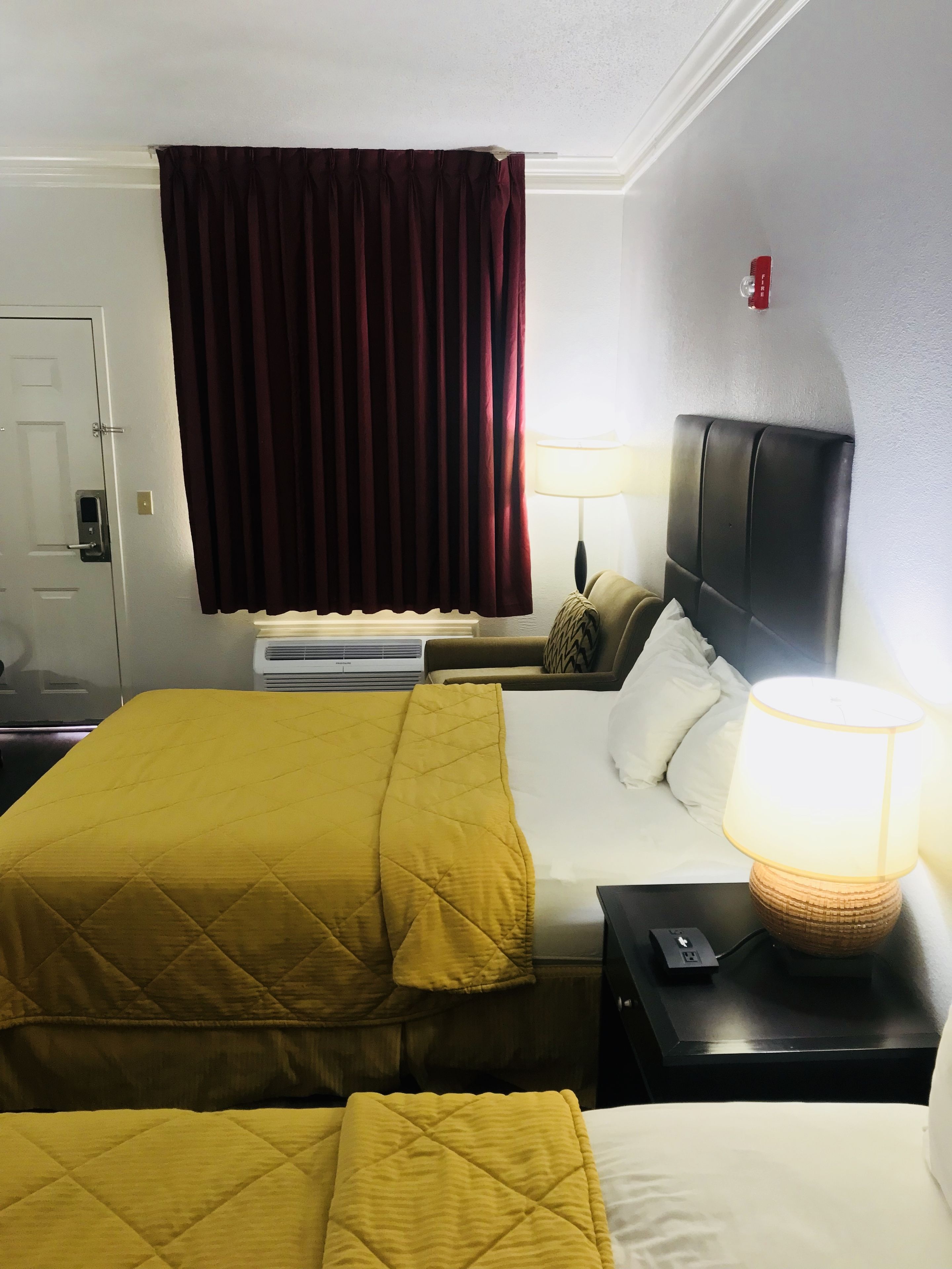 Habitación de hotel con dos camas, un ventanal grande con cortinas oscuras, una lámpara de noche, un televisor y un termostato.