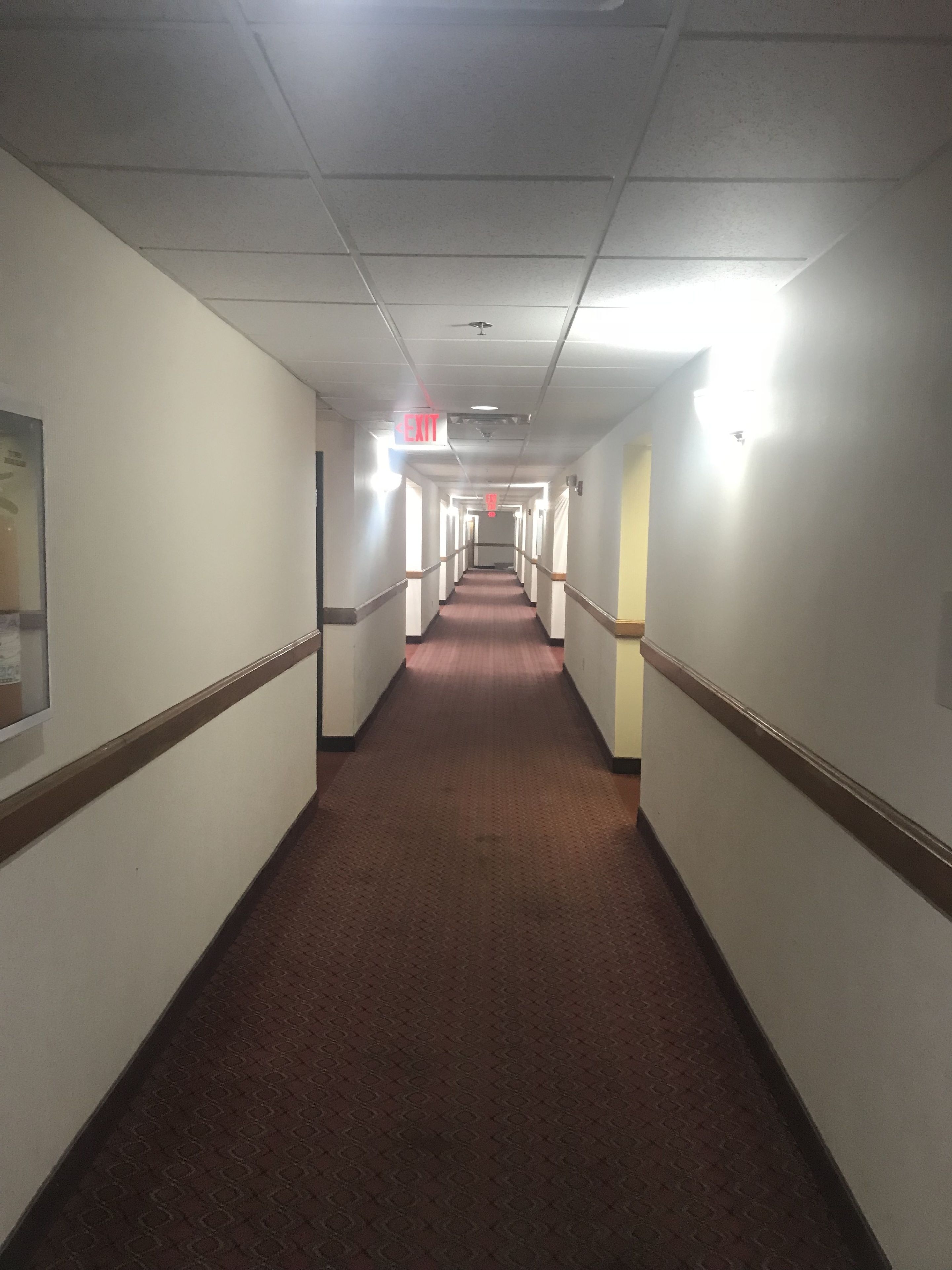 A long, narrow hallway with white walls and a brown carpet. There are doors on both sides, and an exit sign is visible on the left wall.