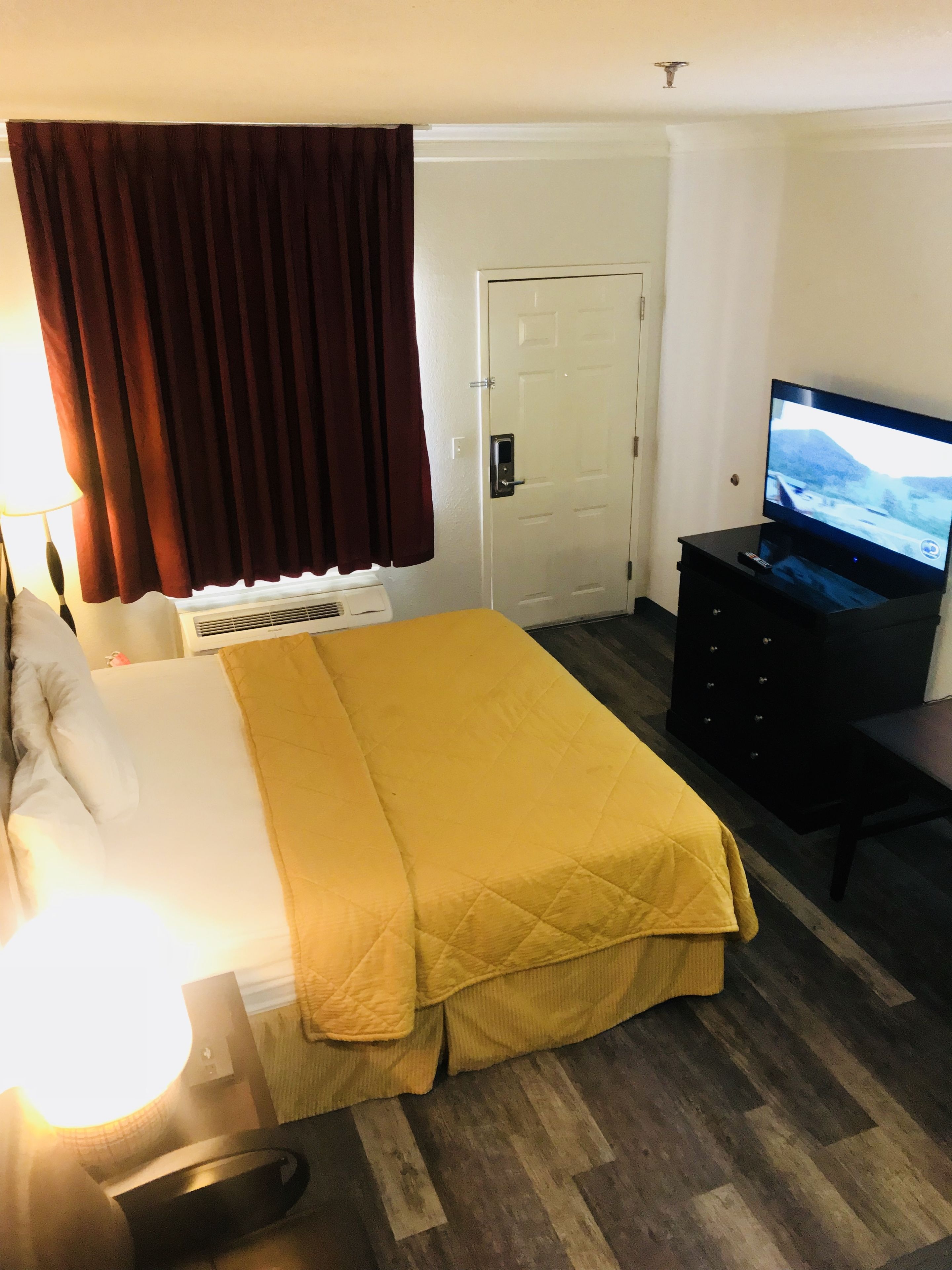 A hotel room with a bed, a television on a stand, a door, and a window with curtains.
