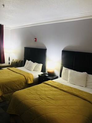 A hotel room with two beds, each with a headboard, a bedside table with a lamp, and a red fire alarm on the wall.