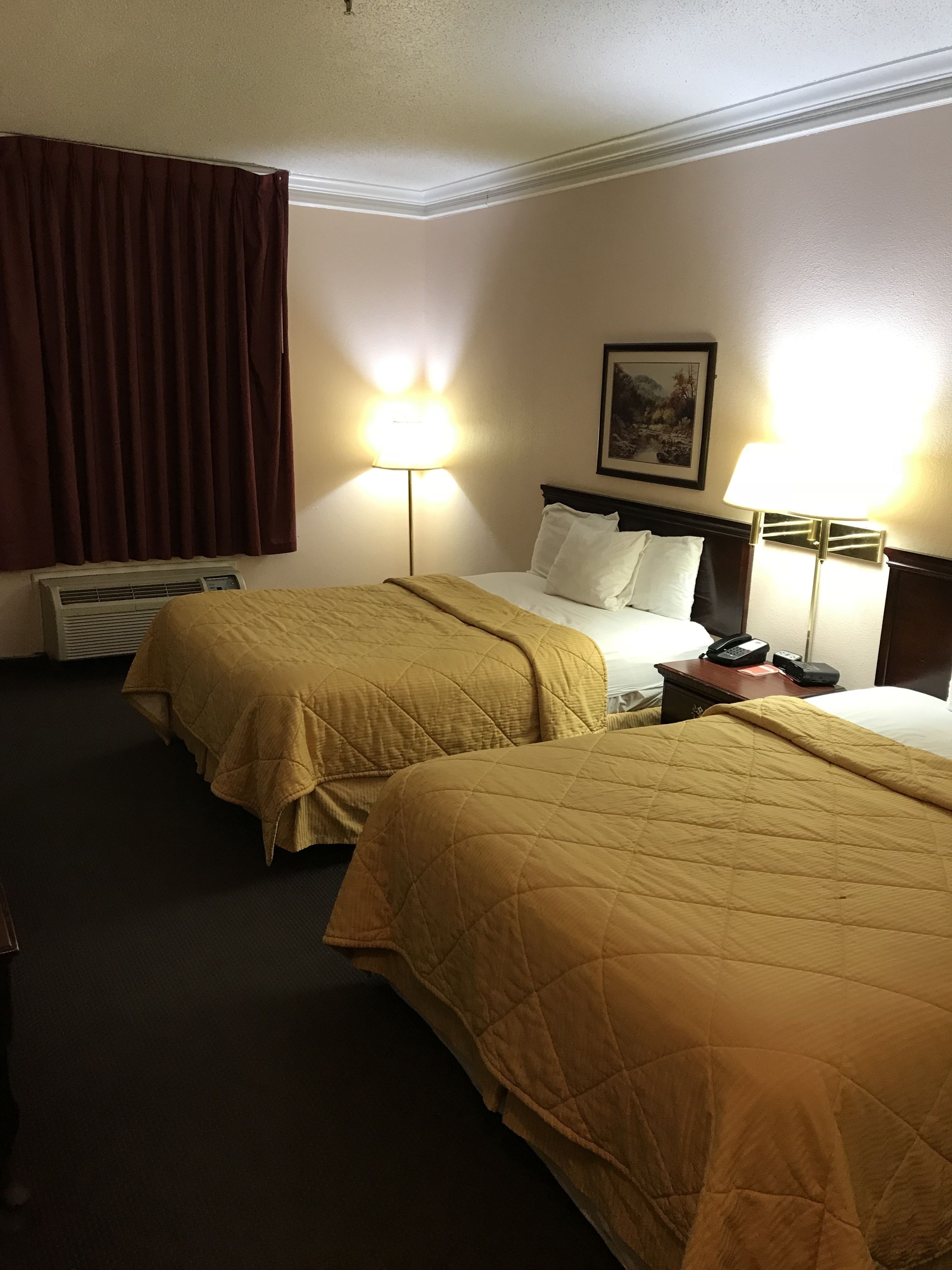 A hotel room with two beds, each with a beige quilted bedspread, a bedside table with a lamp, and a wall-mounted painting.