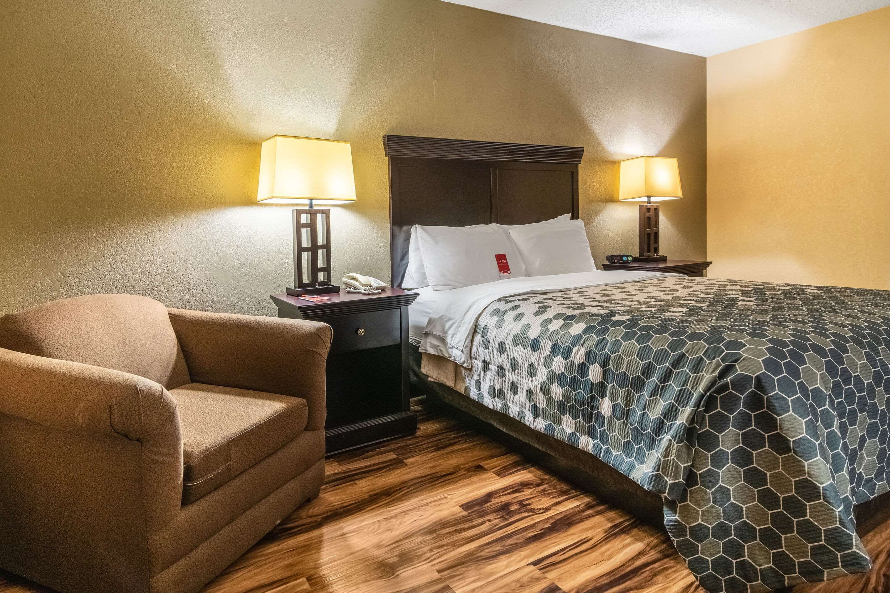 Una habitación de hotel con una cama, dos lámparas de noche, un sillón marrón, suelo de madera y una mesita de noche con un teléfono.