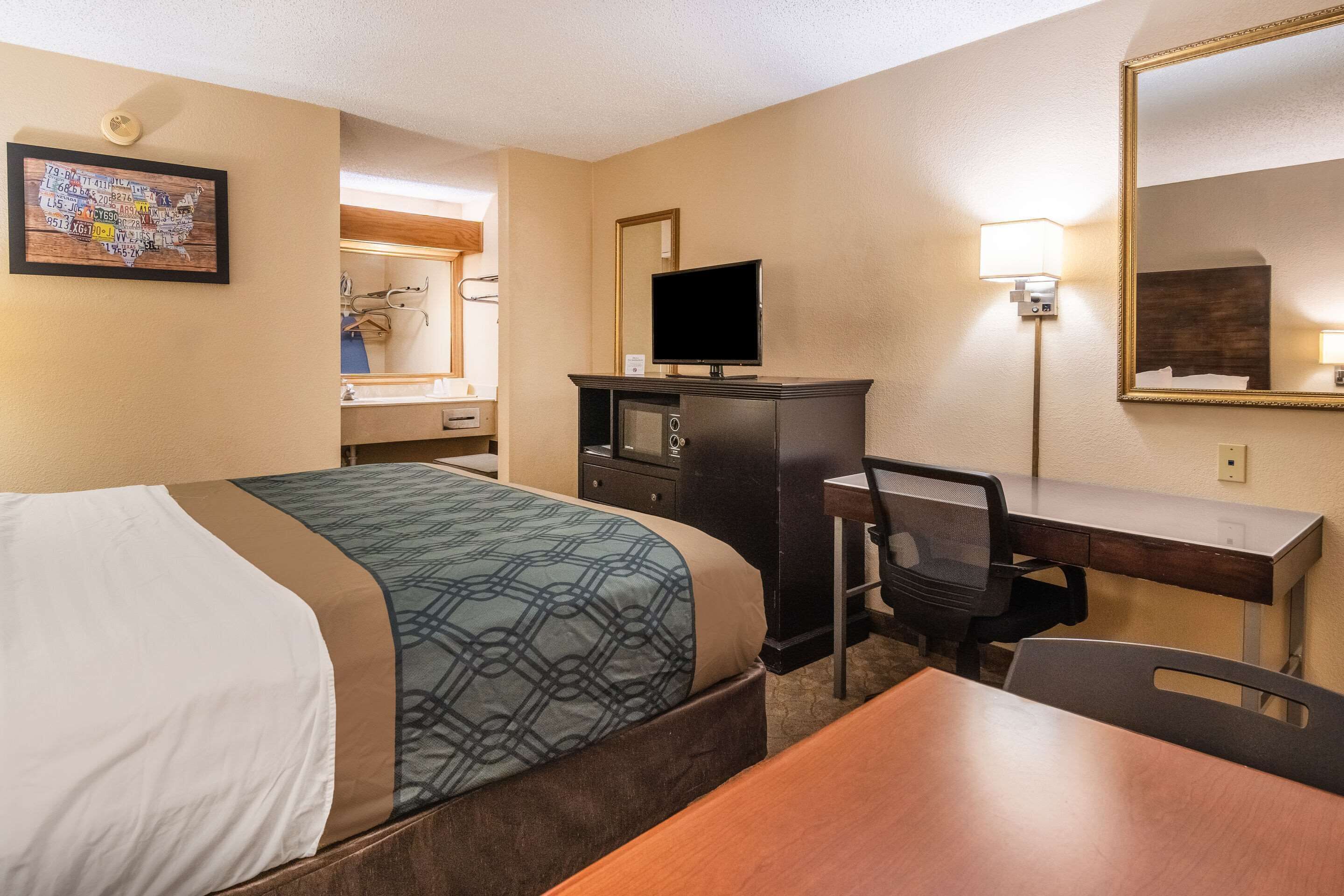 Habitación de hotel con cama, escritorio, silla, televisión, espejo y lámpara de pared.