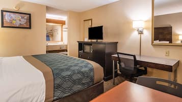Habitación de hotel con cama, escritorio, silla, televisión, espejo y lámpara de pared.