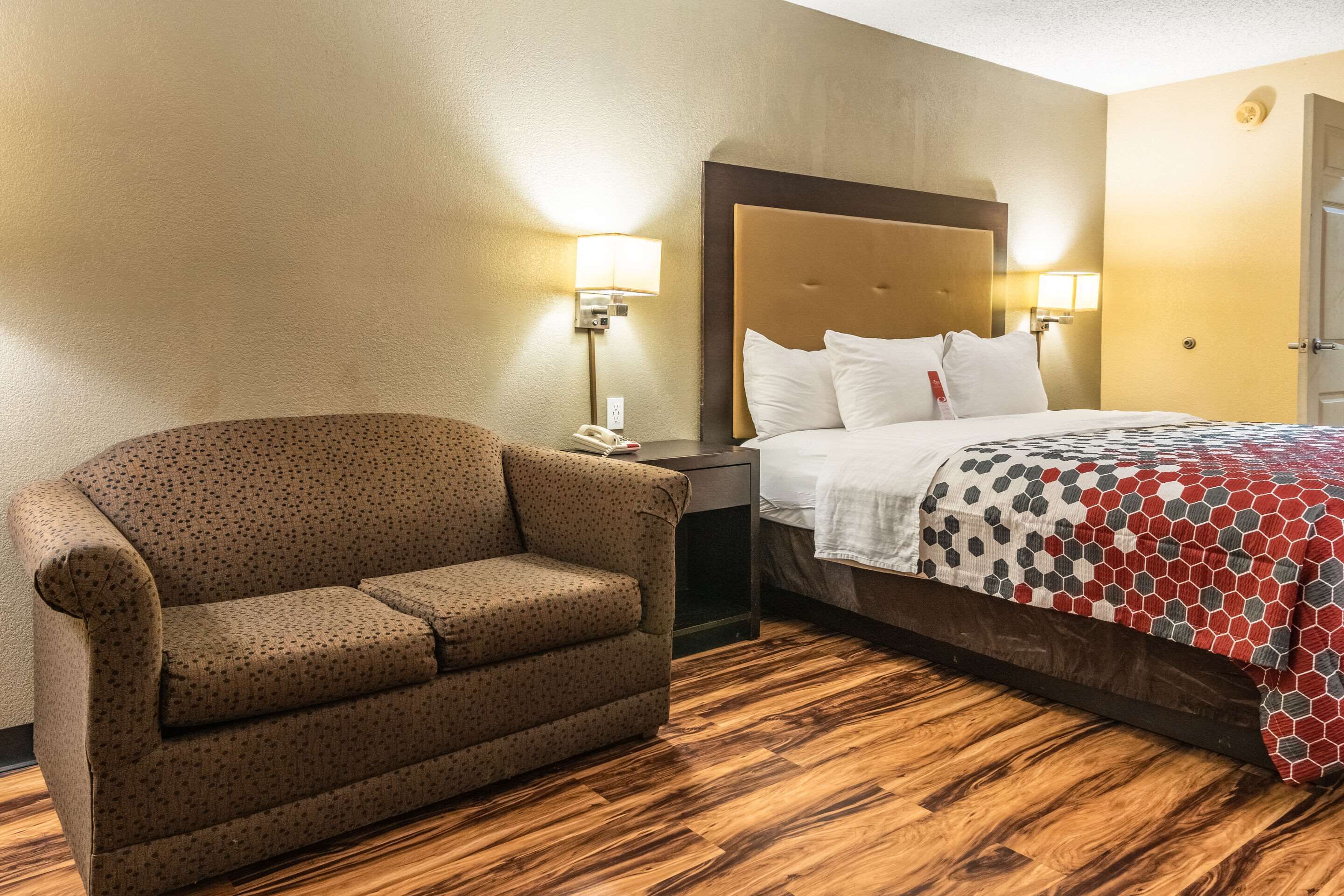A hotel room with a sofa, a bed with a patterned bedspread, wooden flooring, and two wall-mounted lamps.