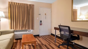 Habitación de hotel con suelo de madera, sofá color beige, mesa de madera, silla de oficina negra, puerta blanca y espejo montado en la pared.
