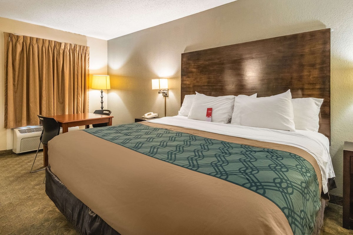 A hotel room with a large bed, a desk with a lamp, and an air conditioning unit.