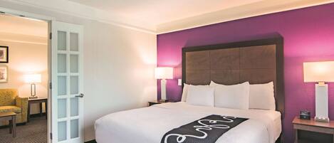 Habitación de hotel con una cama grande, dos mesitas de noche, una silla y una puerta que da a otra habitación.