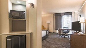 A hotel room with a microwave on a shelf, a bed with a headboard, a desk with a chair, a television, and a window with curtains. - La Quinta Inn & Suites by Wyndham Oklahoma City Norman (Norman)