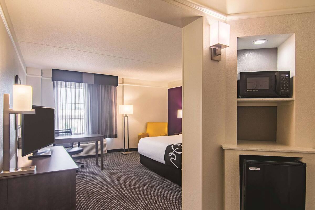 A hotel room with a bed, desk, chair, lamp, and a microwave.