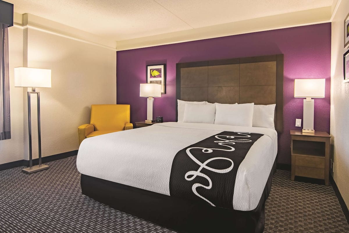 A hotel room with a bed, a chair, two bedside tables with lamps, and a purple accent wall.