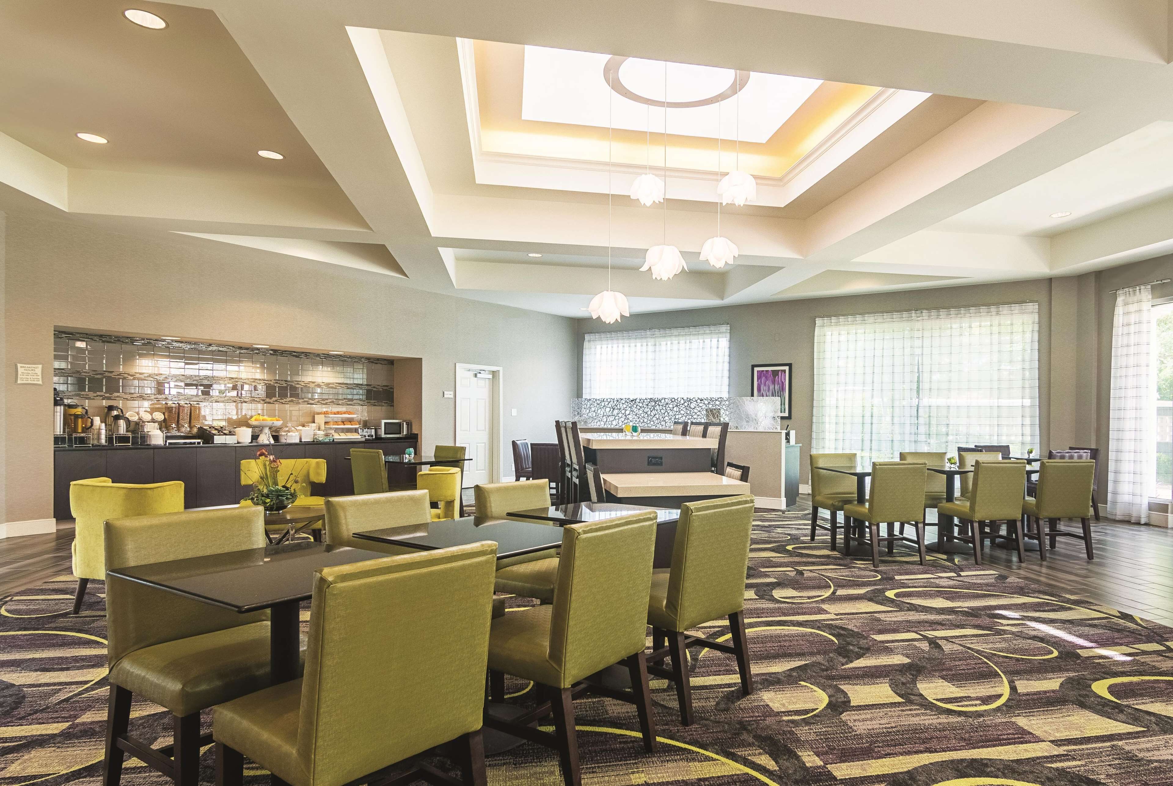 A hotel dining area with a central table and chairs, a kitchen in the background, and a large window with blinds.