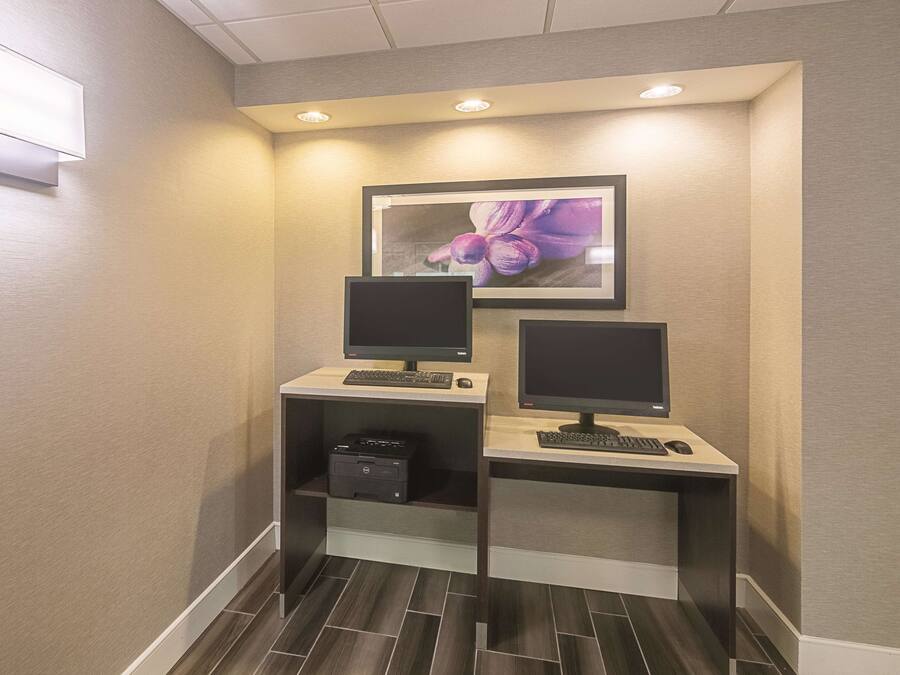 A small office area with two desks, each with a computer monitor, keyboard, and mouse. There is a printer on one of the desks. The walls are beige, and there is a framed picture of flowers on the wall above the desks.
