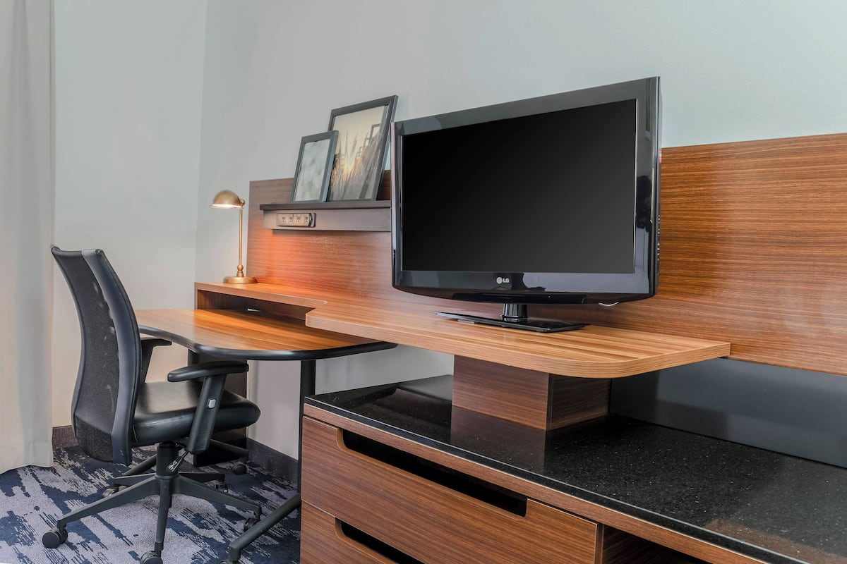 A modern hotel room with a wooden desk, a black office chair, a flat-screen TV, and a small shelf with framed pictures.