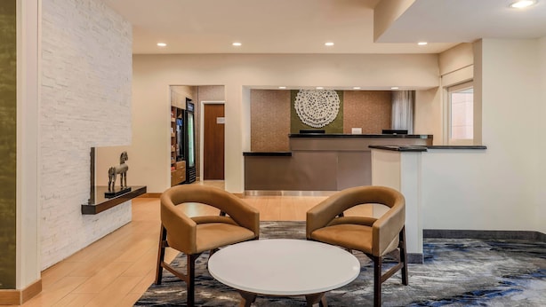 A modern hotel lobby with a reception desk, two leather armchairs, a round white table, and a decorative wall piece.