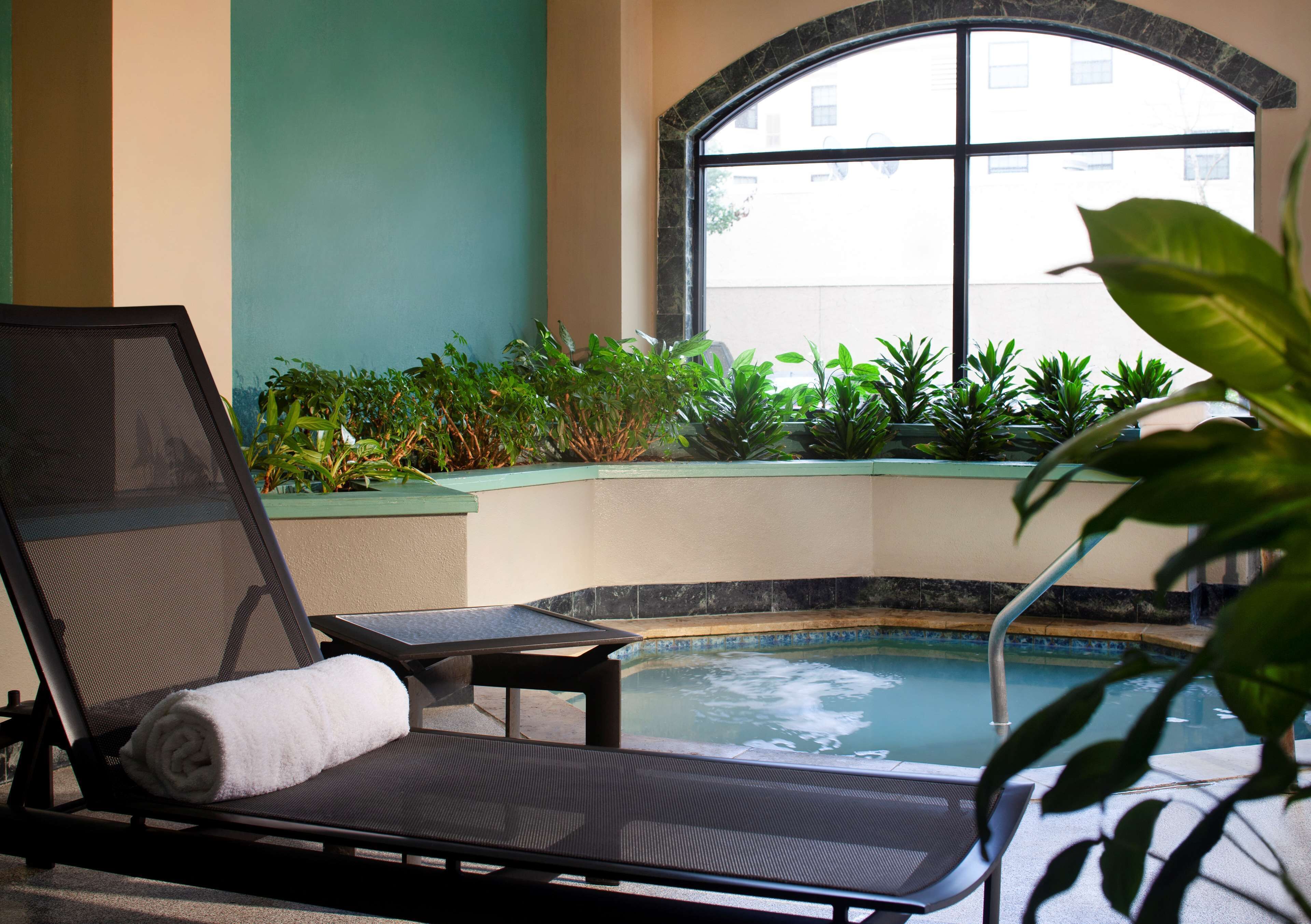 A sunlit indoor pool area with a lounge chair, a small table, and potted plants.