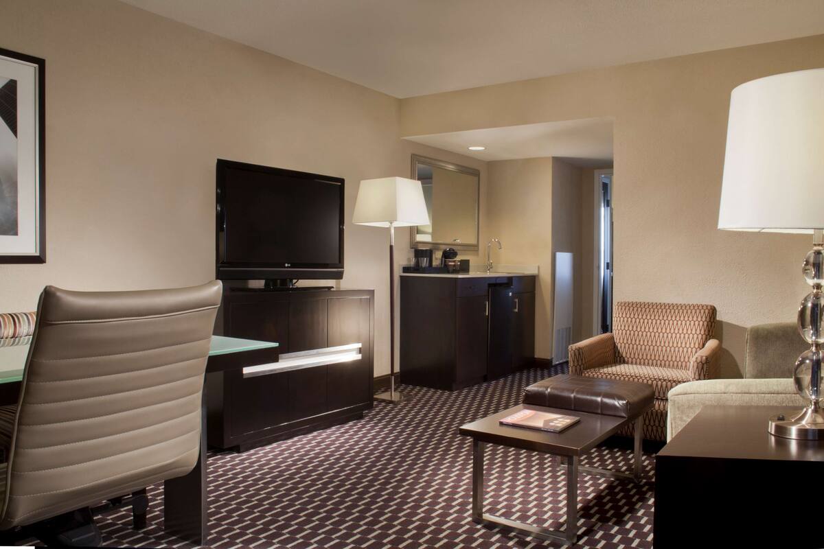 Hotelkamer met flatscreen televisie, bureau met lamp, een bank, salontafel en een kamerbreed tapijt met dessin.