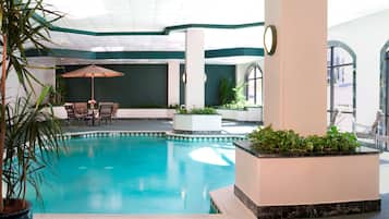 Una piscina interior con borde de azulejos, rodeada de plantas en macetas, una mesa con sillas y un gran paraguas.