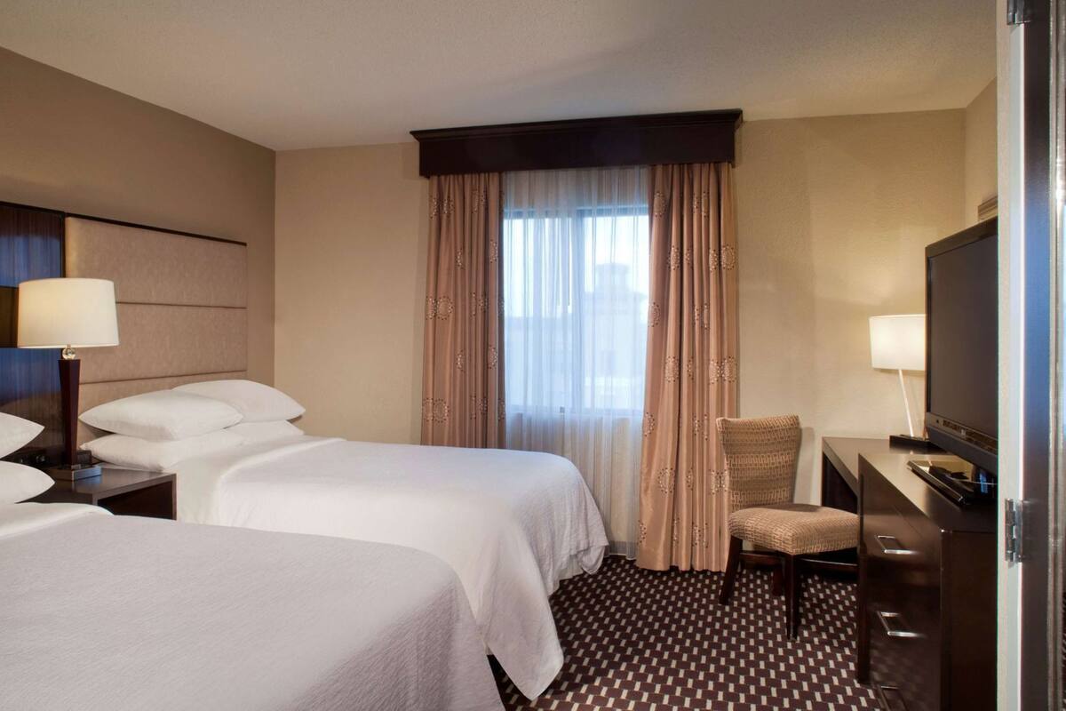 Een hotelkamer met twee bedden, een bureau met een televisie, een stoel en een groot raam met gordijnen.
