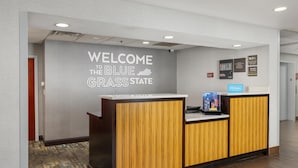 A reception desk with a "Welcome to the Blue Grass State" sign, a Hilton logo, and a digital screen displaying information. - Hampton Inn Carrollton (Carrollton)
