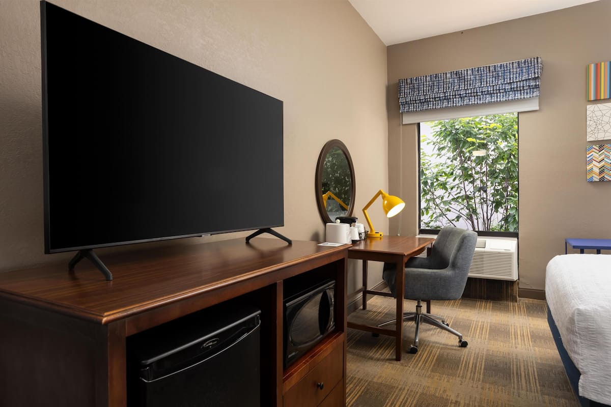 A hotel room with a large flat-screen TV, a wooden desk, a chair, a bed, a window with a view of trees, and a mirror.