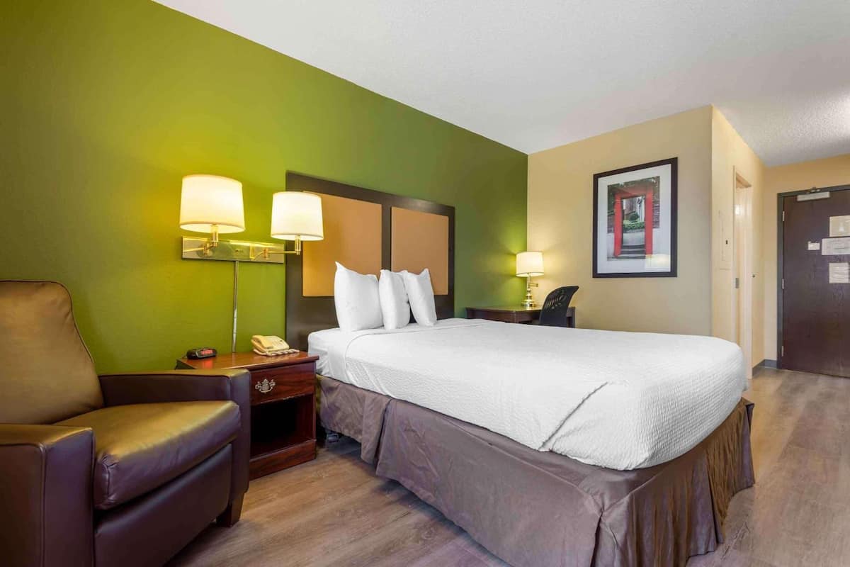 A hotel room with a large bed, a chair, a nightstand with a lamp, and a framed picture on the wall.