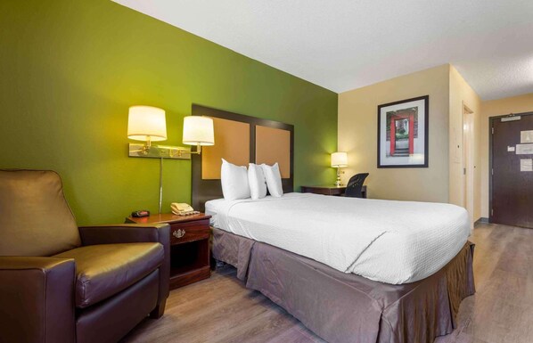 A hotel room with a large bed, a chair, a nightstand with a lamp, and a framed picture on the wall.