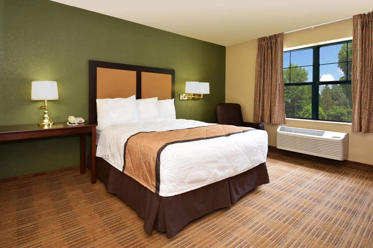 A hotel room with a large bed, two bedside lamps, a desk with a telephone, a chair, and a window with curtains.