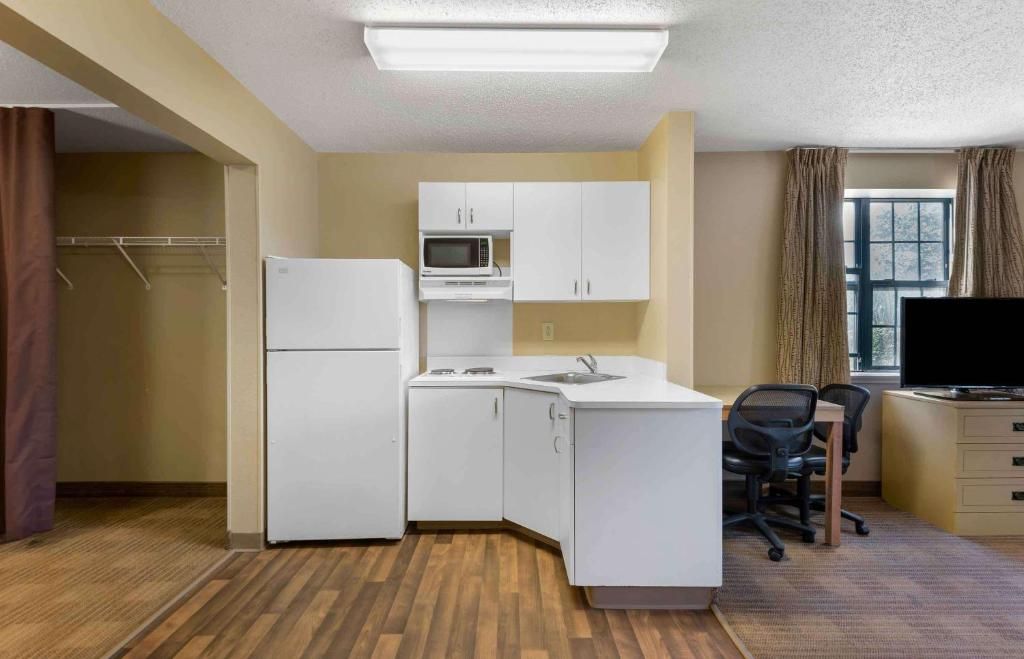 A compact kitchenette with a refrigerator, microwave, and stove, a small desk with a chair, and a window with curtains.